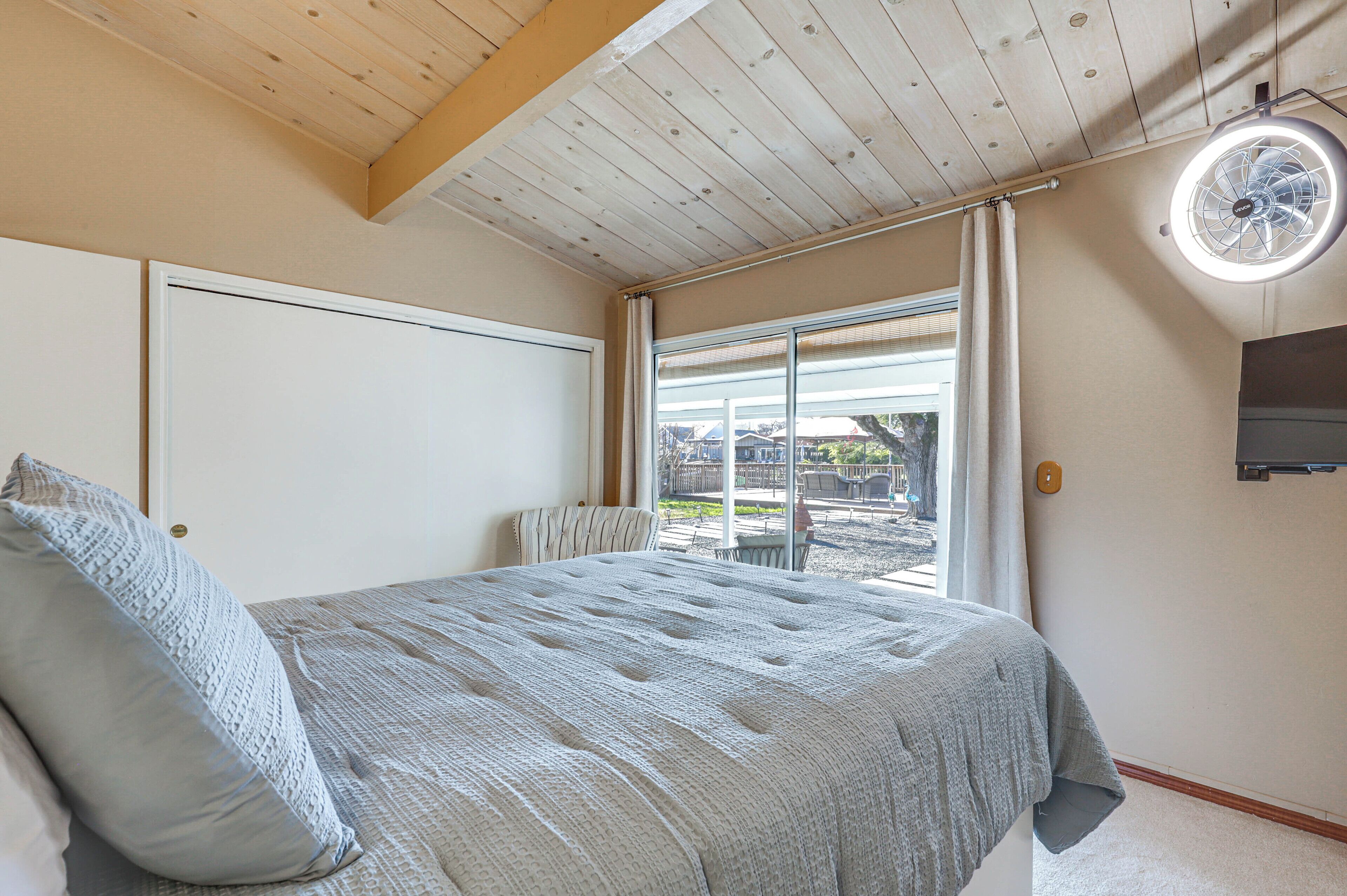 A bedroom with a large bed, a ceiling fan, a sliding glass door to a balcony, and a TV mounted on the wall.