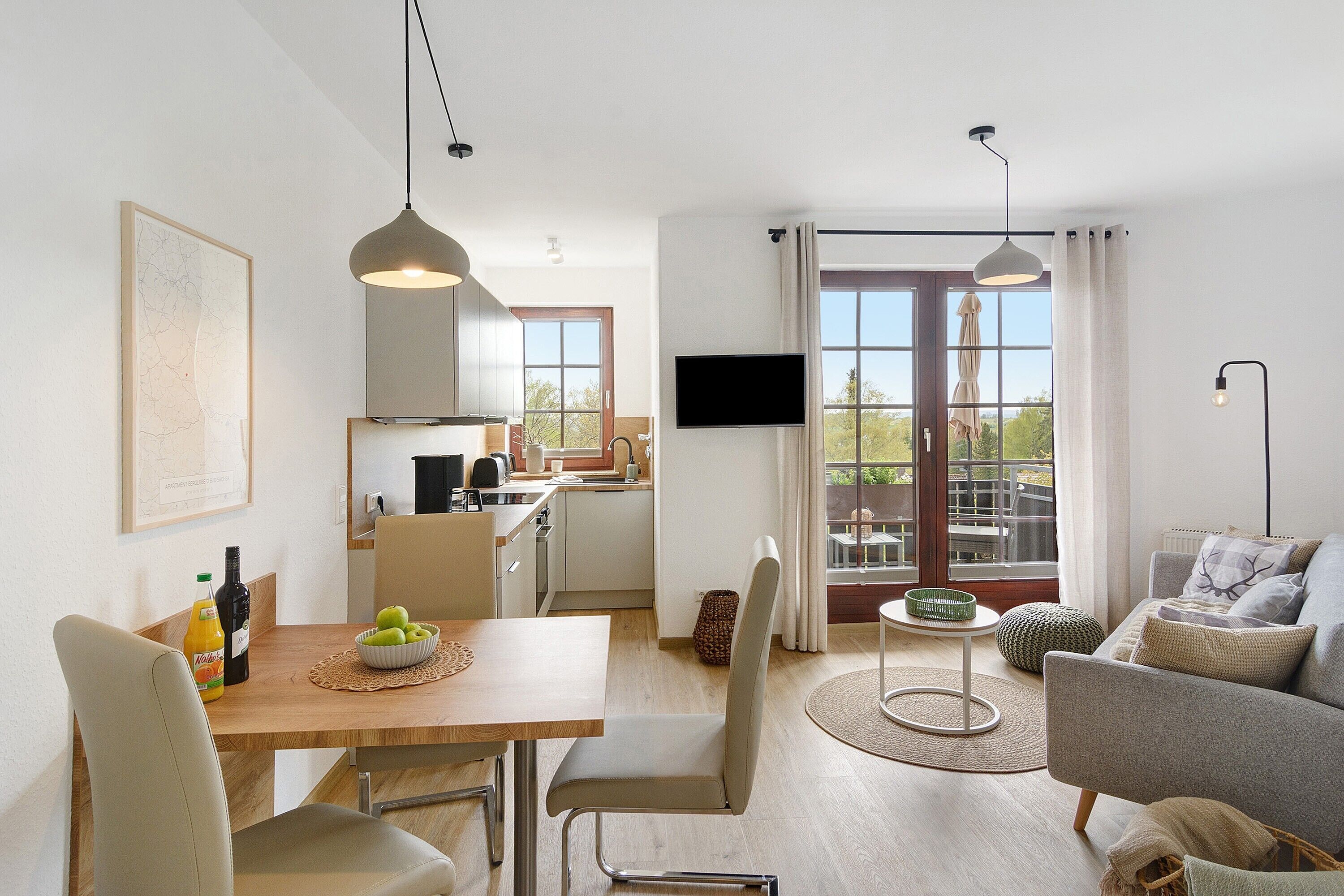 Ein modernes Wohnzimmer mit einer Couch, einem Esstisch mit Stühlen, einer Küchenzeile und einem Balkon mit Fernseher.