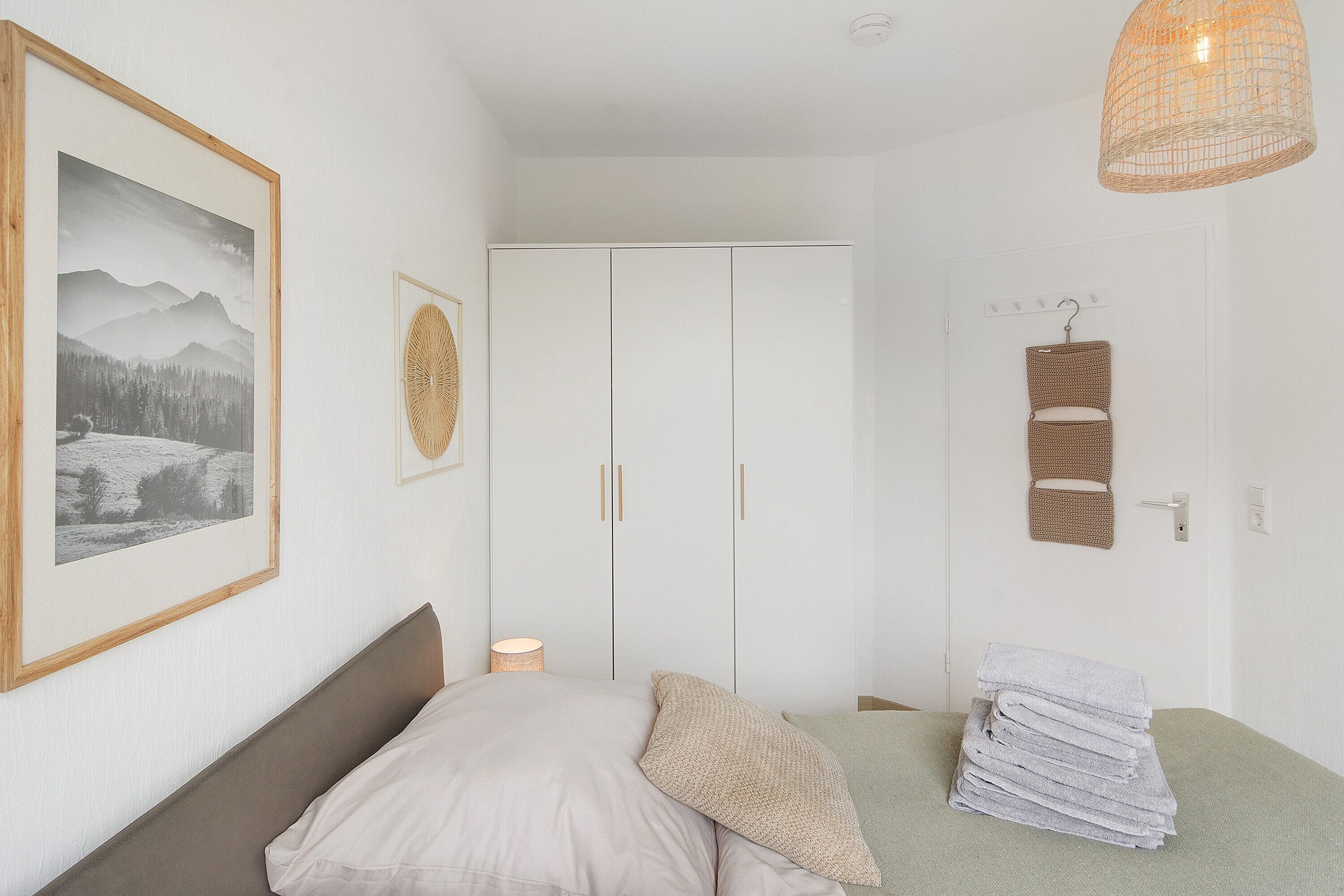 Quarto com cama, guarda-roupa, quadro emoldurado, cesto pendurado e luminária.