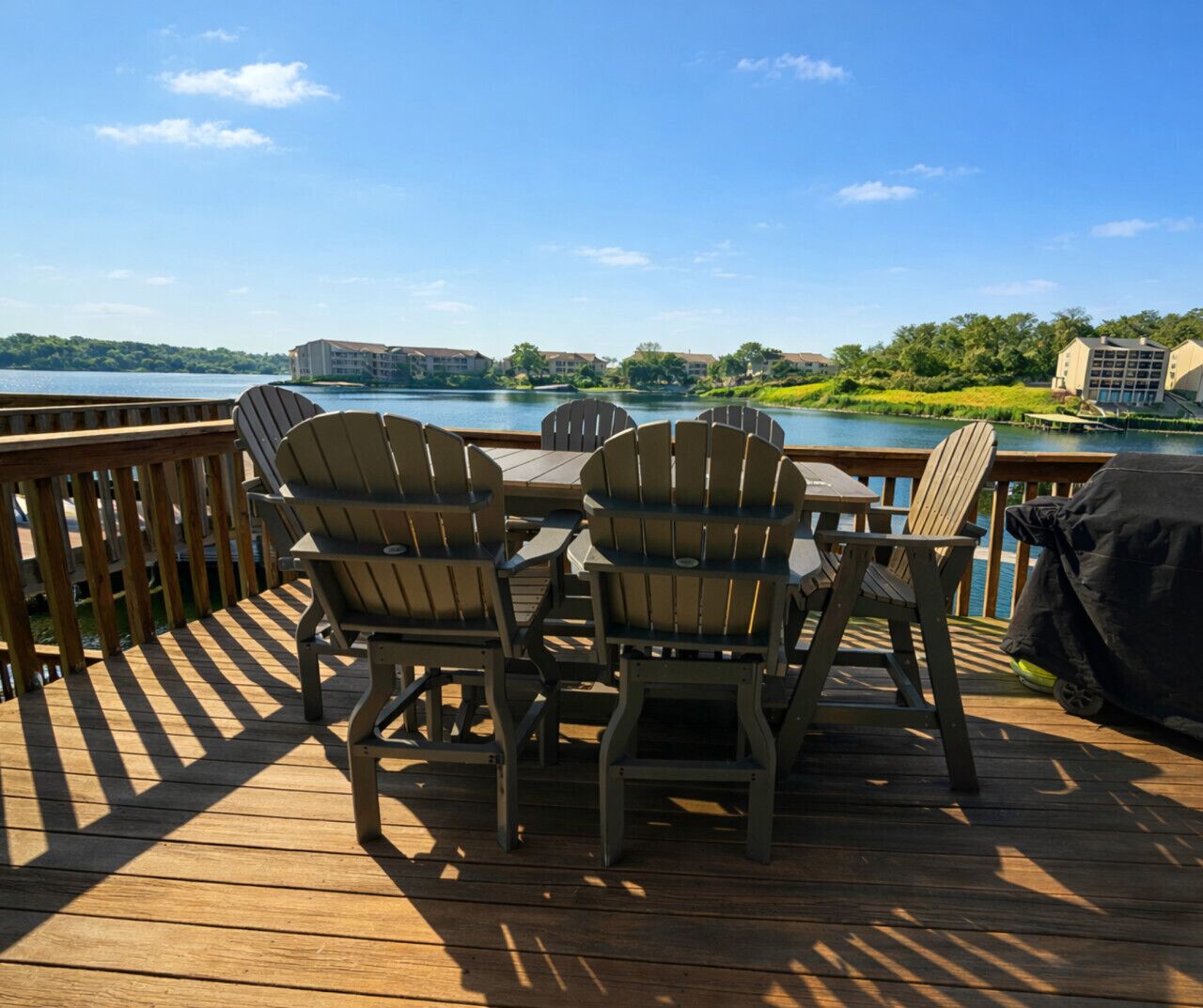 Terraza de madera con sillas Adirondack y una parrilla, con vista a un lago y edificios al fondo.