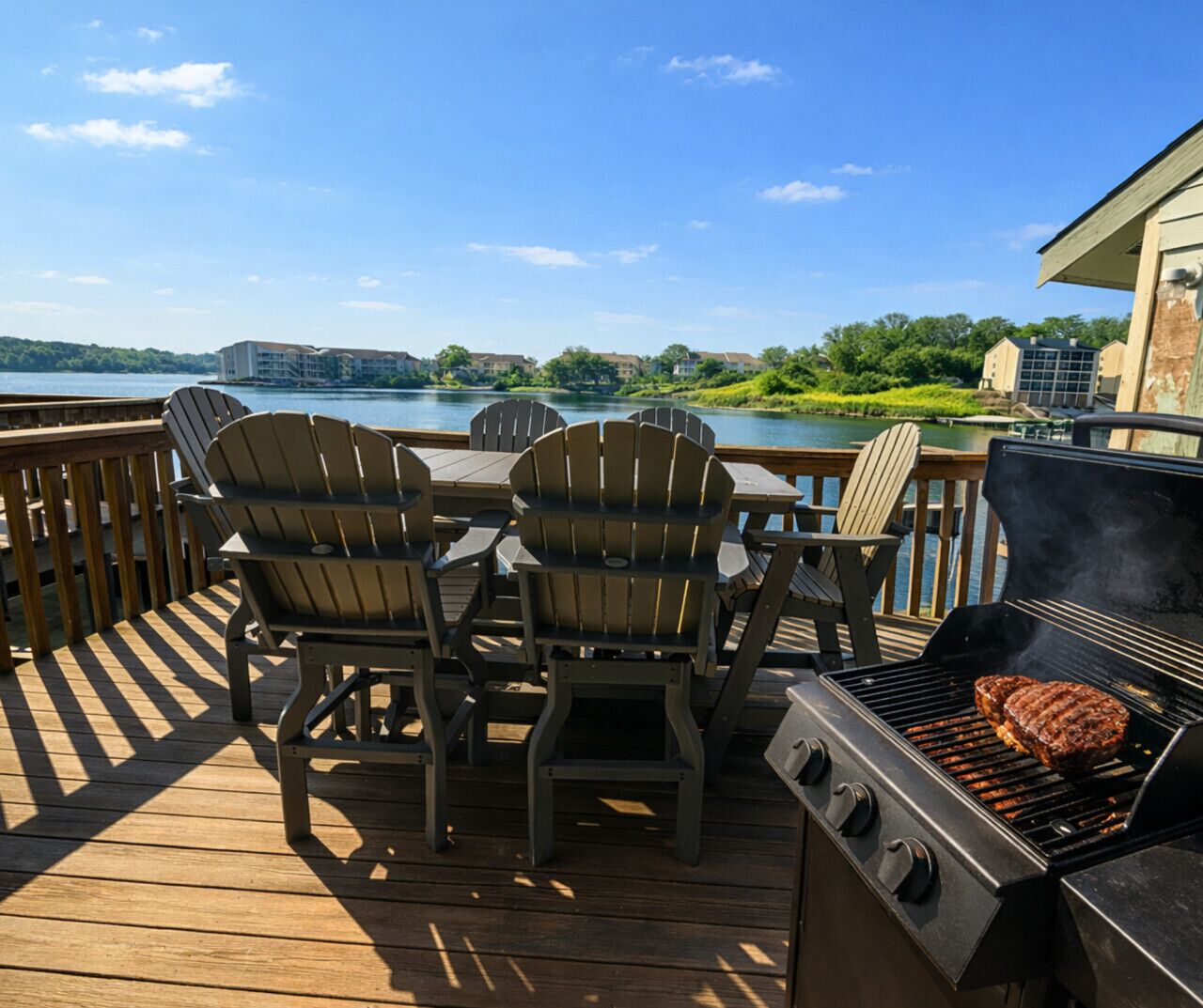 Una terraza con sillas Adirondack y una parrilla, con vista a un lago y edificios al fondo.