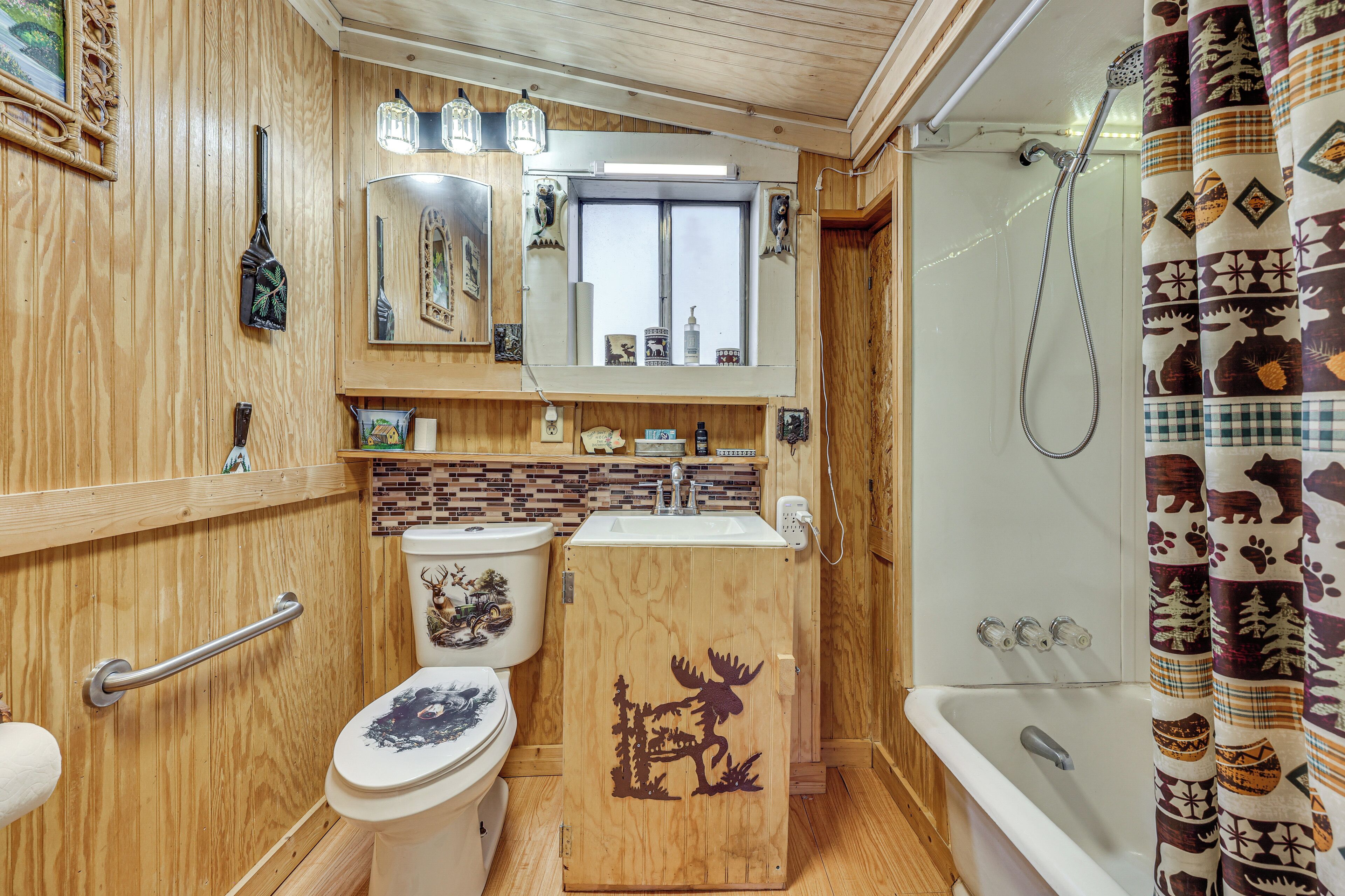 A compact bathroom with wooden walls, a toilet with a moose design, a sink, a mirror, and a shower curtain with animal prints.