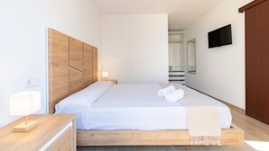 Ein modernes Hotelzimmer mit einem Holzkopfteil, einem Bett mit weißen Leinen, einem Nachttisch mit Lampe, einem an der Wand montierten Flachbildfernseher und einem Holzschrank.