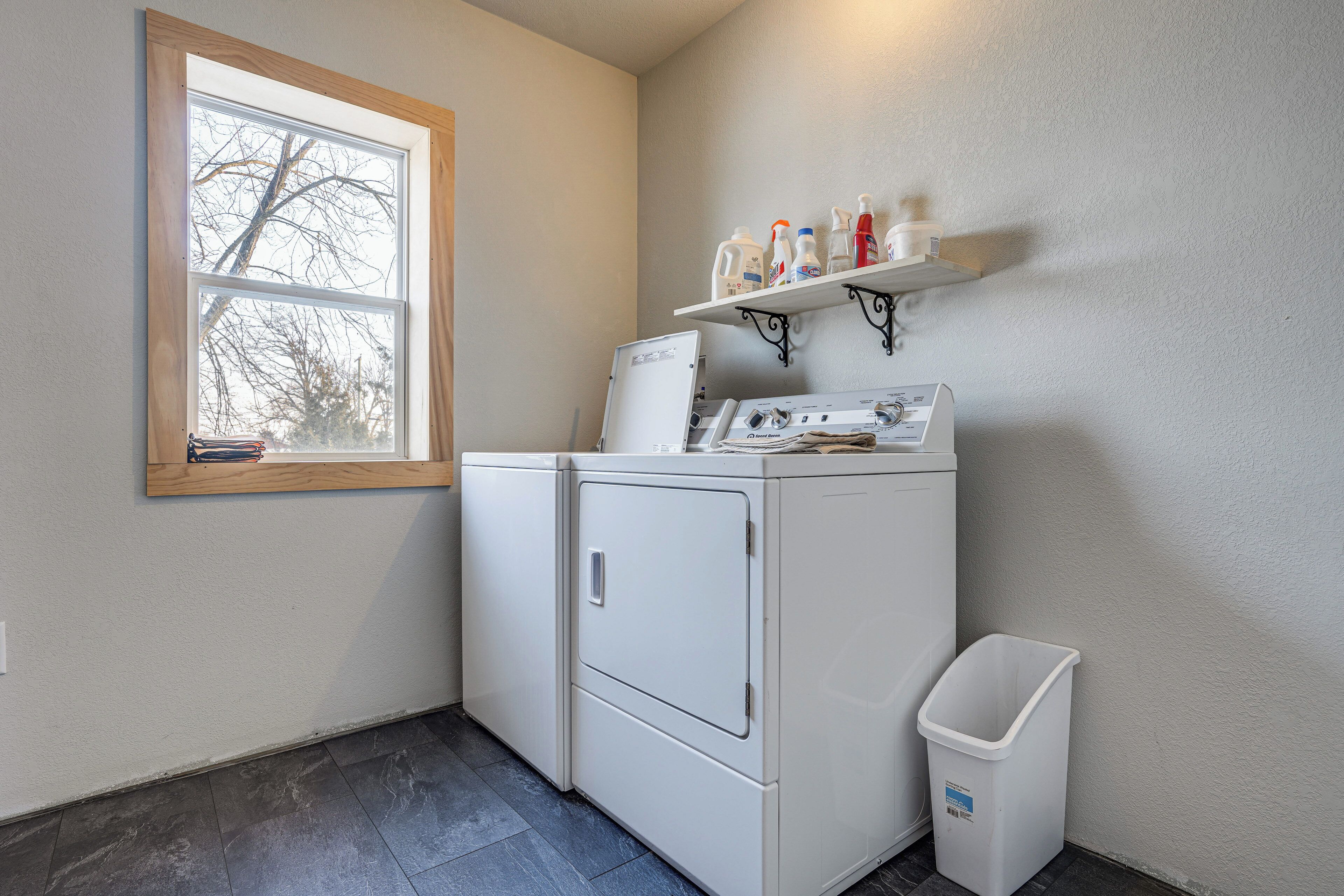 A laundry room with a washer and dryer, a window, a shelf with cleaning supplies, and a trash bin.