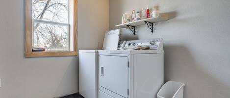 A laundry room with a washer and dryer, a window, a shelf with cleaning supplies, and a trash bin.