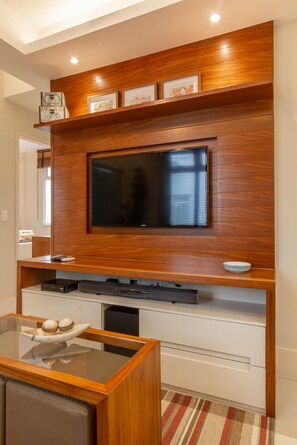 Een moderne woonkamer met een grote flatscreen-tv aan een houten wand, een glazen salontafel met decoratieve objecten en een witte tv-meubel eronder.