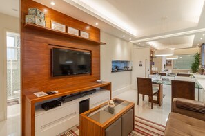 Een moderne woonkamer met een flatscreen-televisie, een houten tv-meubel, een glazen salontafel en een eethoek met stoelen en een glazen tafel.