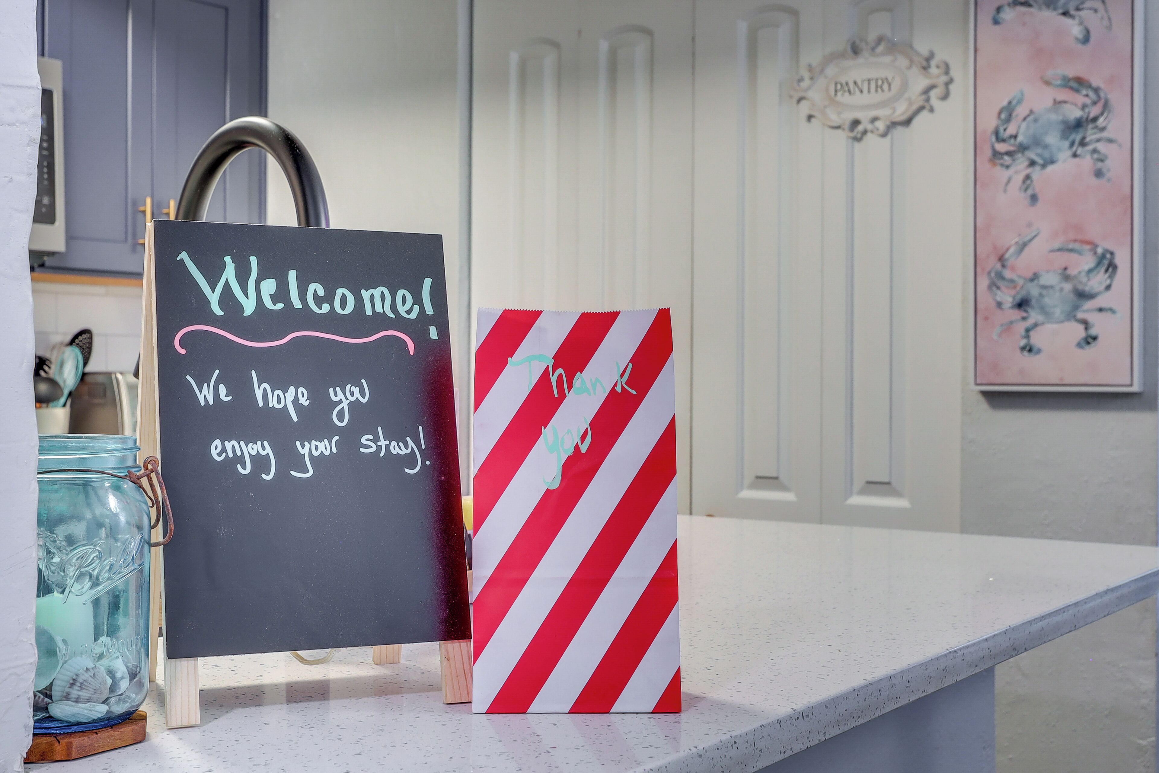 A kitchen counter with a chalkboard sign that reads "Welcome! We hope you enjoy your stay!" and a red and white striped bag with "Thank You" written on it.
