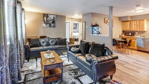 A living room with a sofa set, coffee table, and a rug. There is a kitchen area with wooden cabinets and a dining table. - Easily Explore Anchorage! Modern Duplex Near Town (Anchorage)
