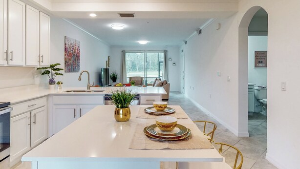 A modern kitchen with white cabinetry, a central island with gold-rimmed bowls, and a potted plant. - Play & Stay: 1st Floor Ava Maria Condo! (Immokalee)