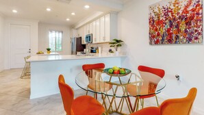 A modern kitchen with white cabinets, a central island, and a dining area with orange chairs and a glass table. - Play & Stay: 1st Floor Ava Maria Condo! (Immokalee)