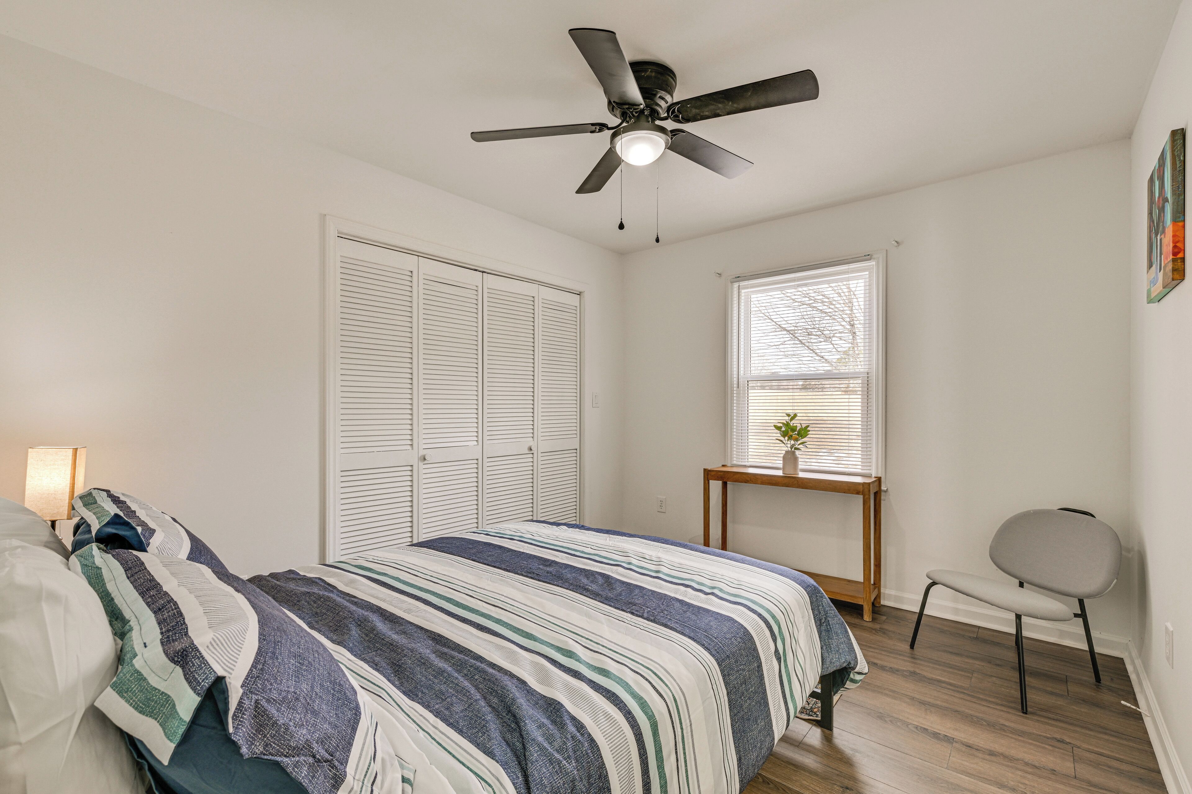 A bedroom with a bed, a ceiling fan, a window, a chair, and a small table with a plant.