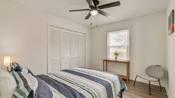 A bedroom with a bed, a ceiling fan, a window, a chair, and a small table with a plant.