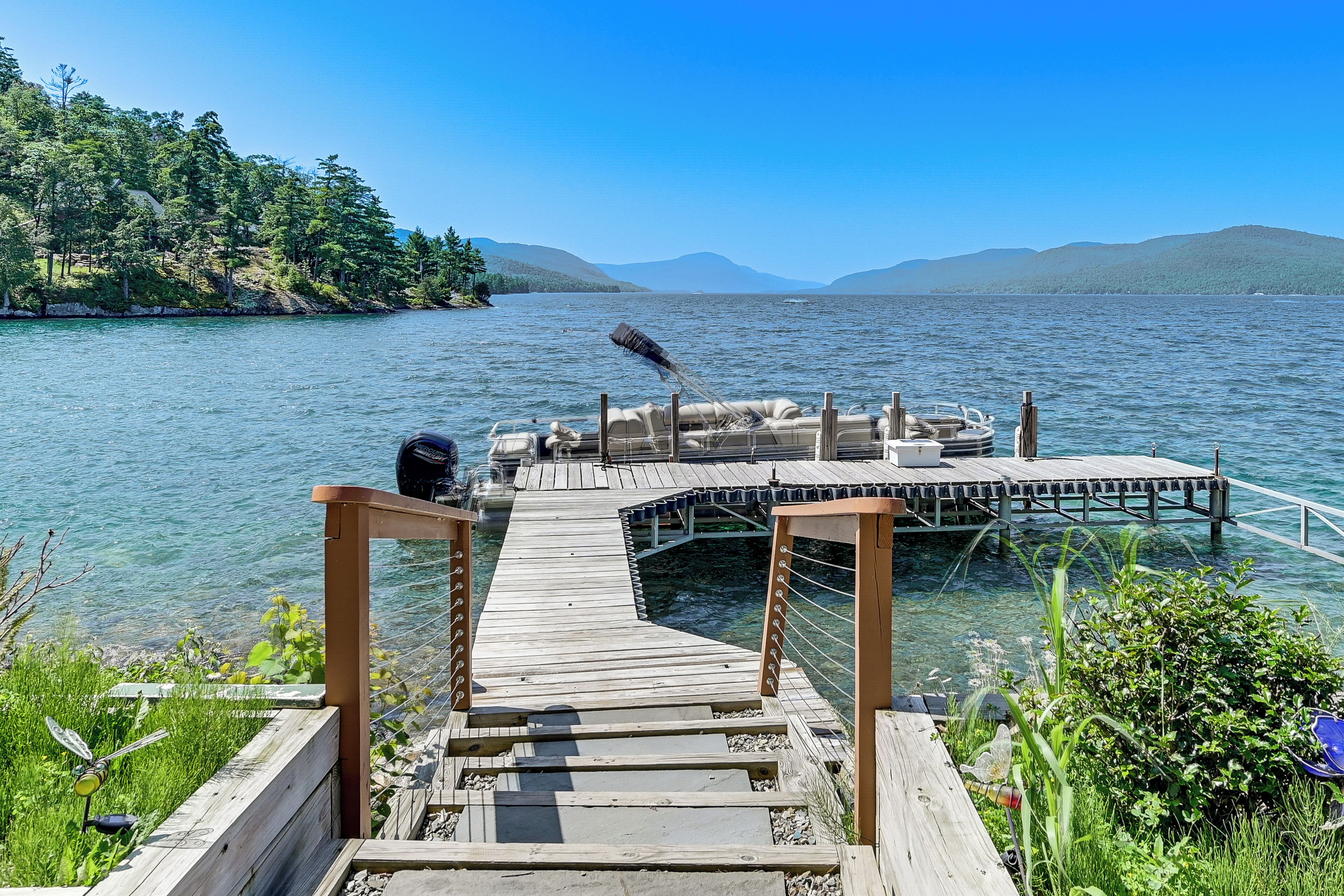 A wooden dock extending into a lake with clear water, surrounded by greenery and mountains in the background.