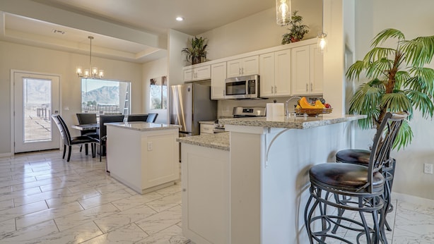 A modern kitchen with white cabinets, granite countertops, and stainless steel appliances. There are two bar stools and a dining area with a table and chairs. - Mountain-view Retreat Near Organ Wilderness Trails (Las Cruces)
