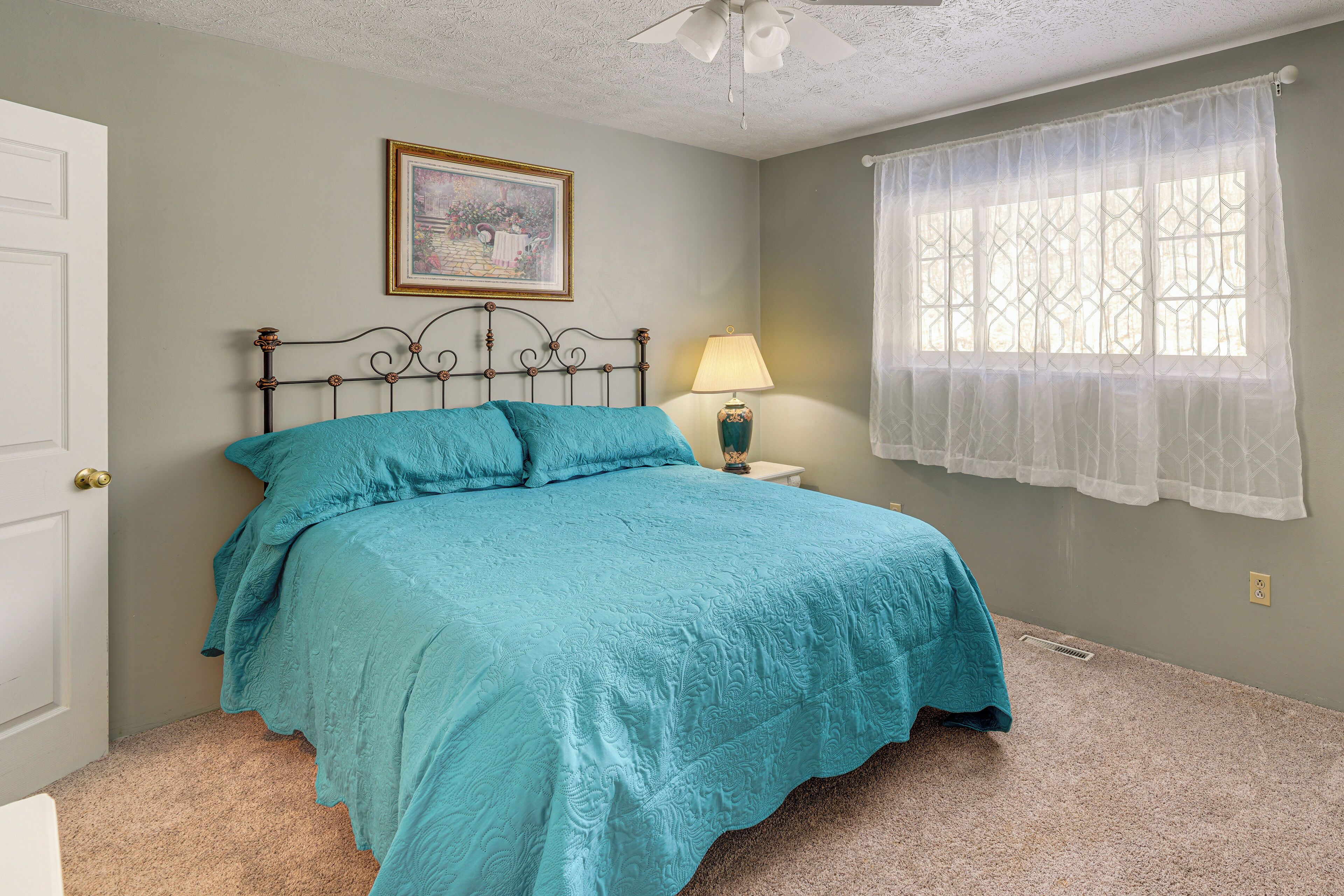 A bedroom with a bed, a nightstand, a lamp, a window with sheer curtains, and a ceiling fan.