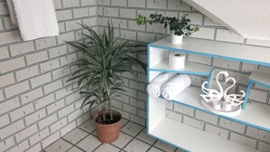 Een badkamer met een betegelde muur, een plant in pot, een witte handdoek op een rek en een blauwe plank met toiletartikelen.