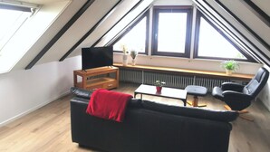 Een moderne loft slaapkamer met een schuin dak, een houten tv-meubel, een zwarte leren bank, een rode plaid, een kleine tafel met een plant, en een zwarte fauteuil.