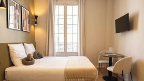 Una camera d'albergo con un letto, un tavolino con sedie, una TV a muro e una finestra con tende.