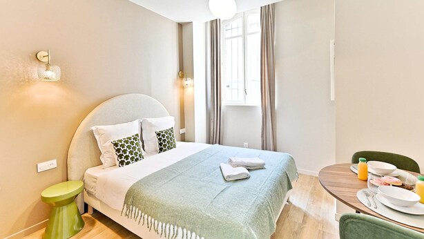 A hotel room with a bed, bedside table, lamp, curtains, window, and breakfast table with plates, bowls, and juice. - Studio 15m² avec clim – Pratique et fonctionnel (Le Pré-Saint-Gervais)