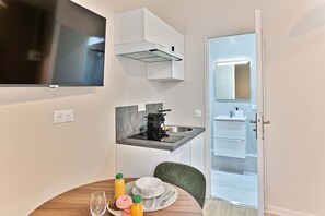 A compact kitchen with a built-in stove, a small sink, and a microwave. There is a dining table with a chair, and a door leading to a bathroom. - Studio 15m² avec clim – Pratique et fonctionnel (Le Pré-Saint-Gervais)