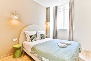 A neatly made bed with a green throw, a bedside lamp, and a window with curtains. - Studio 15m² avec clim – Pratique et fonctionnel (Le Pré-Saint-Gervais)