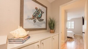 Un pasillo con una consola blanca, una obra de arte con tortuga marina enmarcada, una planta en maceta y vistas a un dormitorio con cama y ventana.