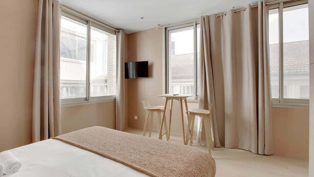Quarto de hotel com cama, televisão fixada na parede, mesinha e duas cadeiras, e janelas amplas com cortinas.