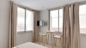 A hotel room with a bed, a TV, a small table, two chairs, and large windows with curtains. - Cocon climatisé confortable – Neuilly (Neuilly-sur-Seine)