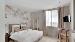 A modern bedroom with a large bed, a bedside table, a TV, and a window with curtains. - Studio climatisé lumineux et cosy (Neuilly-sur-Seine)