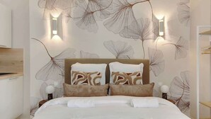 A neatly made bed with a white duvet and brown pillows, a wooden nightstand with a lamp, and a wall with a leaf pattern and two wall-mounted lights. - Studio climatisé lumineux et cosy (Neuilly-sur-Seine)