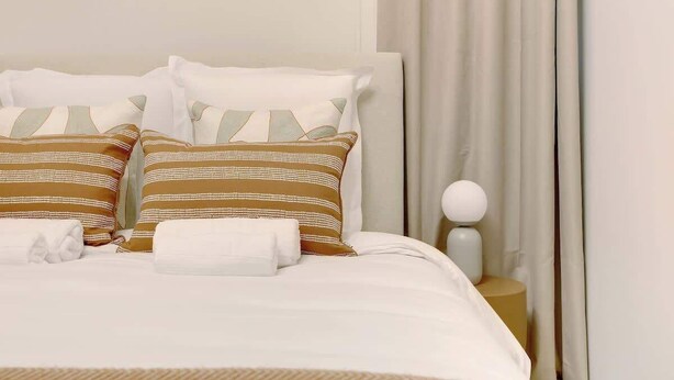 A neatly made bed with striped pillows and a white blanket, a bedside table with a lamp, and a curtain partially drawn. - Air-conditioned studio offering comfort and elegance (Neuilly-sur-Seine)