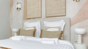 Una cama bien hecha con sábanas blancas y almohadones estampados, dos lámparas fijadas a la pared y dos cuadros enmarcados.
