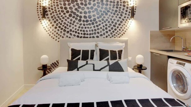 A modern hotel room with a neatly made bed, a washing machine, and a kitchen area. - Studio climatisé intimiste – Neuilly (Neuilly-sur-Seine)