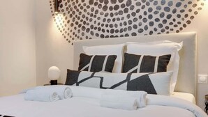 A neatly made bed with white linens and a geometric-patterned blanket, a headboard with multiple pillows, and a wall with a circular dot pattern. - Studio climatisé intimiste – Neuilly (Neuilly-sur-Seine)