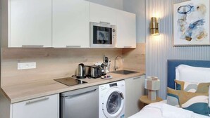A compact hotel room with a kitchenette, including a microwave, toaster, and washing machine. There is a bed with a blue headboard and a bedside table with a lamp. - Studio climatisé confort & charme – Neuilly (Neuilly-sur-Seine)