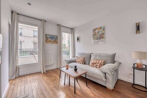 Ein Wohnzimmer mit grauem Sofa, einem Holztisch und einer Lampe. Zwei Fenster mit Vorhängen, ein Heizkörper und ein Bild an der Wand sind ebenfalls vorhanden.