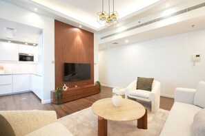A modern living room with a flat-screen TV, a wooden entertainment unit, and a kitchen area with white cabinetry and a built-in oven. - Inviting 1BR at Opal Tower (Dubai)