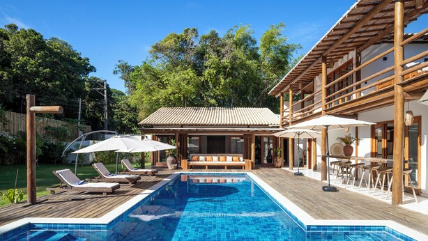 A swimming pool with lounge chairs and umbrellas, wooden deck, and a building with a tiled roof. - Casa Villamar25 (Porto Seguro)