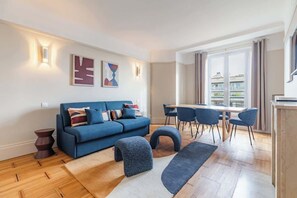 A modern living room with a blue sofa, a dining table with chairs, and a large window with curtains. - Élégant appartement 3 pièces avec balcon – Neuilly-sur-Seine (Neuilly-sur-Seine)