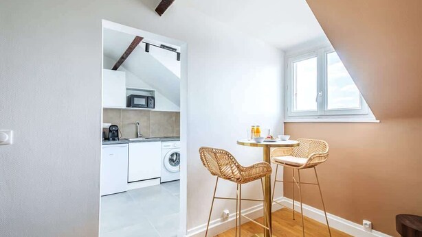 A modern kitchen with white cabinets, a washing machine, and a coffee maker. There is a dining area with two wicker chairs and a small round table. - Studio fonctionnel et calme au cœur d'Issy (Issy-les-Moulineaux)