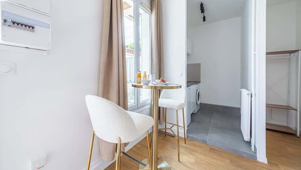 A modern kitchen with a built-in oven, a dining area with a round table and two white chairs, and a view of the outside through a window. - Studio Chic & Cosy au Cœur d'Issy (Issy-les-Moulineaux)