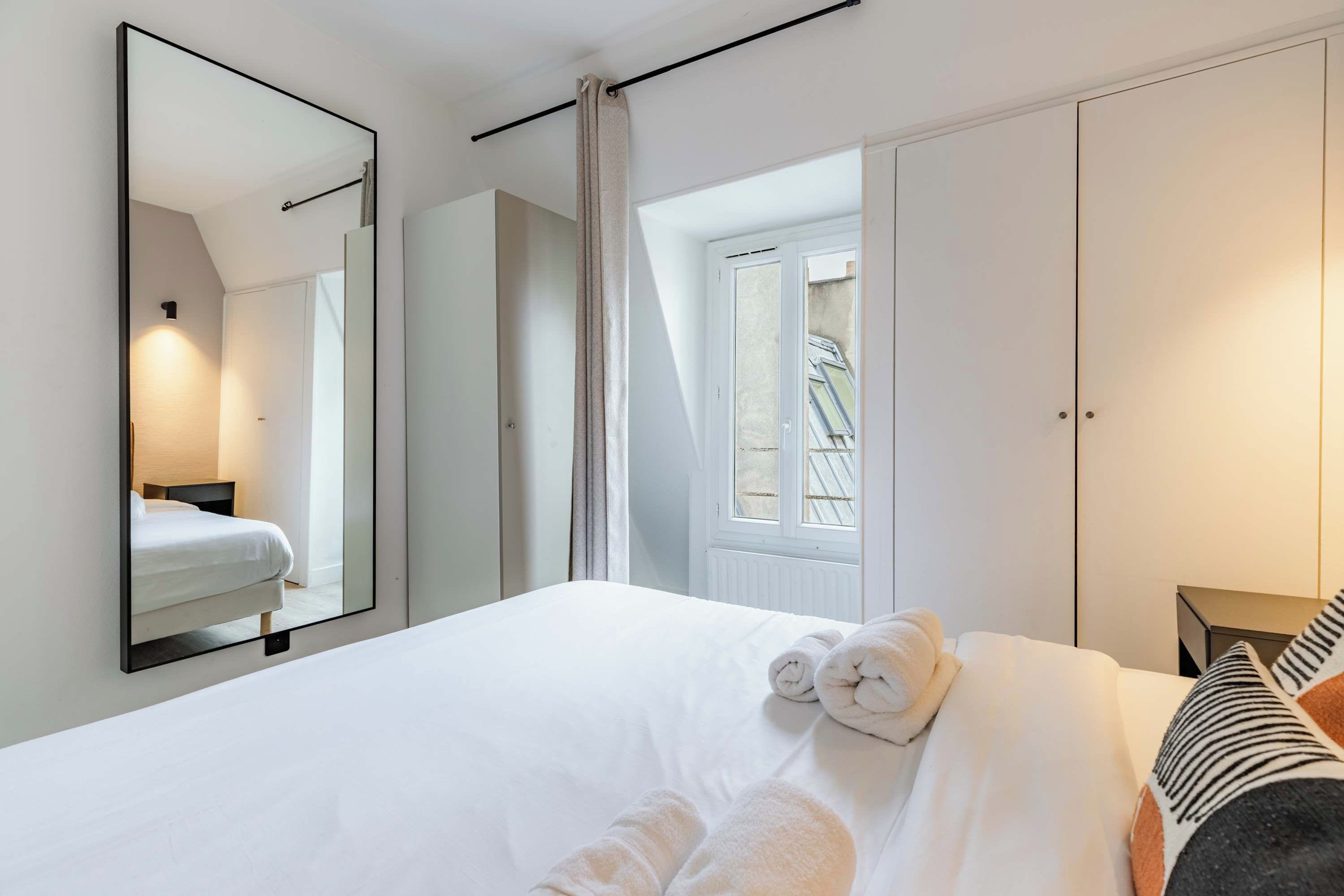 A hotel room with a bed, a large mirror, a wardrobe, a window with curtains, and a nightstand.