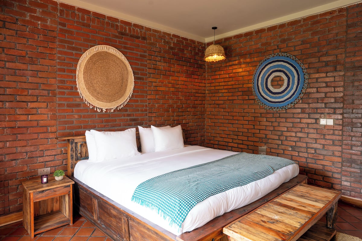 A room with a brick wall, a bed with white bedding, a wooden bench, a small table with a candle, and two wall decorations.