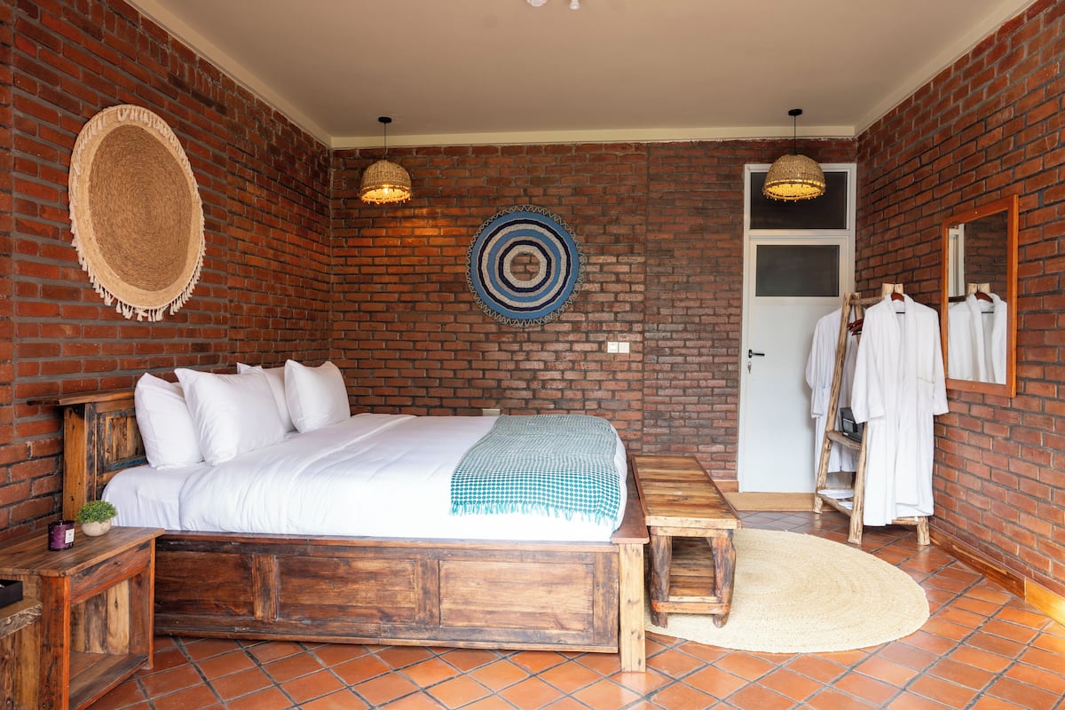 A room with a brick wall, a bed with white bedding, a wooden bench, and a small table.