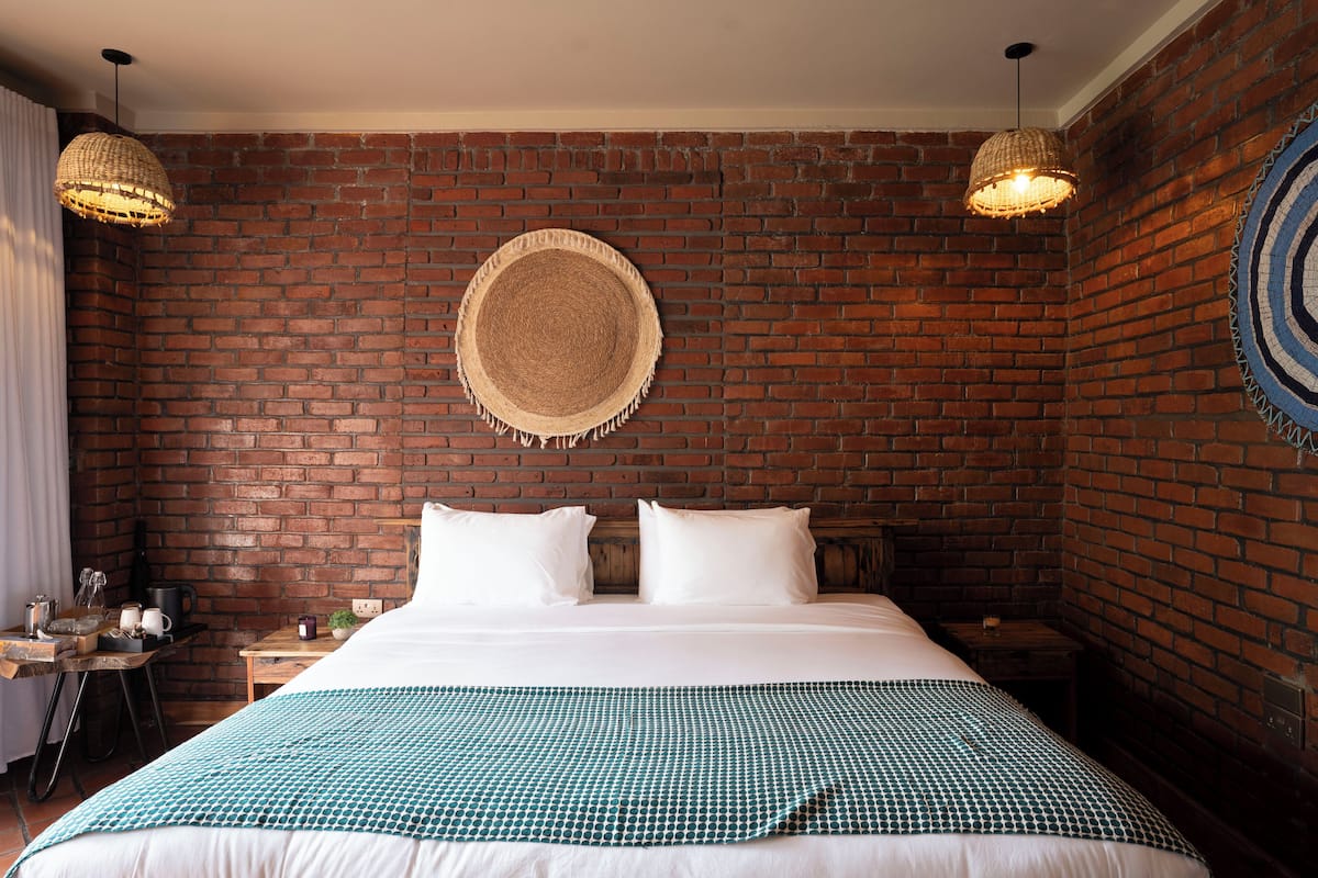 A hotel room with a brick wall, a bed with white linens, two bedside tables, and a woven wall hanging.