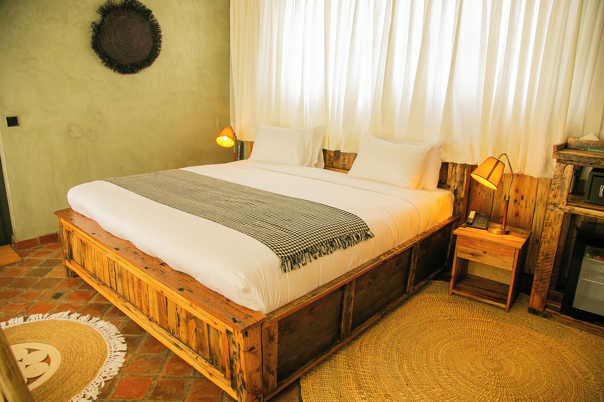 A wooden bed with a white and patterned bedspread, a bedside lamp, and a small wooden nightstand with a telephone.