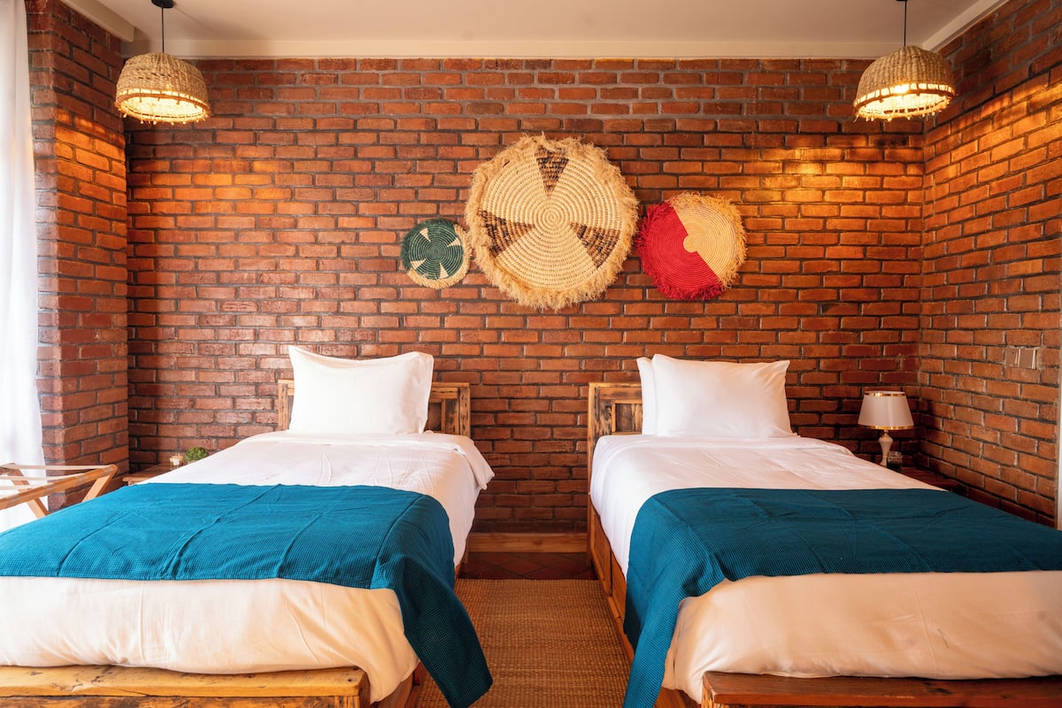 A room with two beds, a brick wall, and woven wall hangings.