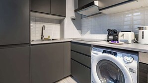 A modern kitchen with grey cabinets, a built-in dishwasher, a washing machine, a coffee maker, and a toaster. - Studio Lumineux au Cœur de Paris Centre (Paris)