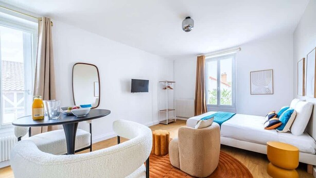 A modern hotel room with a white sofa, a round coffee table, a small round table with a bottle and glasses, a mirror, a TV, and a view of the outside. - Studio chaleureux au coeur de ville Issy (Issy-les-Moulineaux)